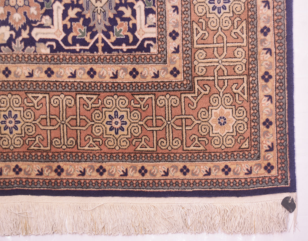 A close-up of a traditional Hereke rug corner showing a peach and beige central field with navy blue edge, cream fringe, and a small dark tag at the bottom right.