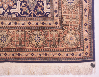 A close-up of a traditional Hereke rug corner showing a peach and beige central field with navy blue edge, cream fringe, and a small dark tag at the bottom right.