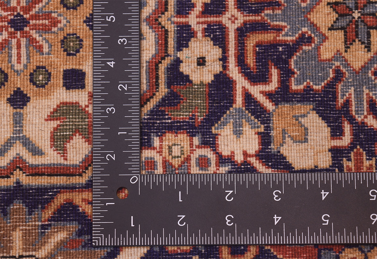 Close-up of the back of a Hereke rug corner with a dark metal ruler placed to show scale, featuring a beige, navy, red, and green ornamental design.