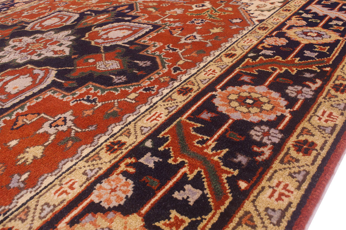 A close-up of a traditional patterned Handmade Indo Heriz carpet with a warm rust red field, dark navy borders, and beige floral motifs.