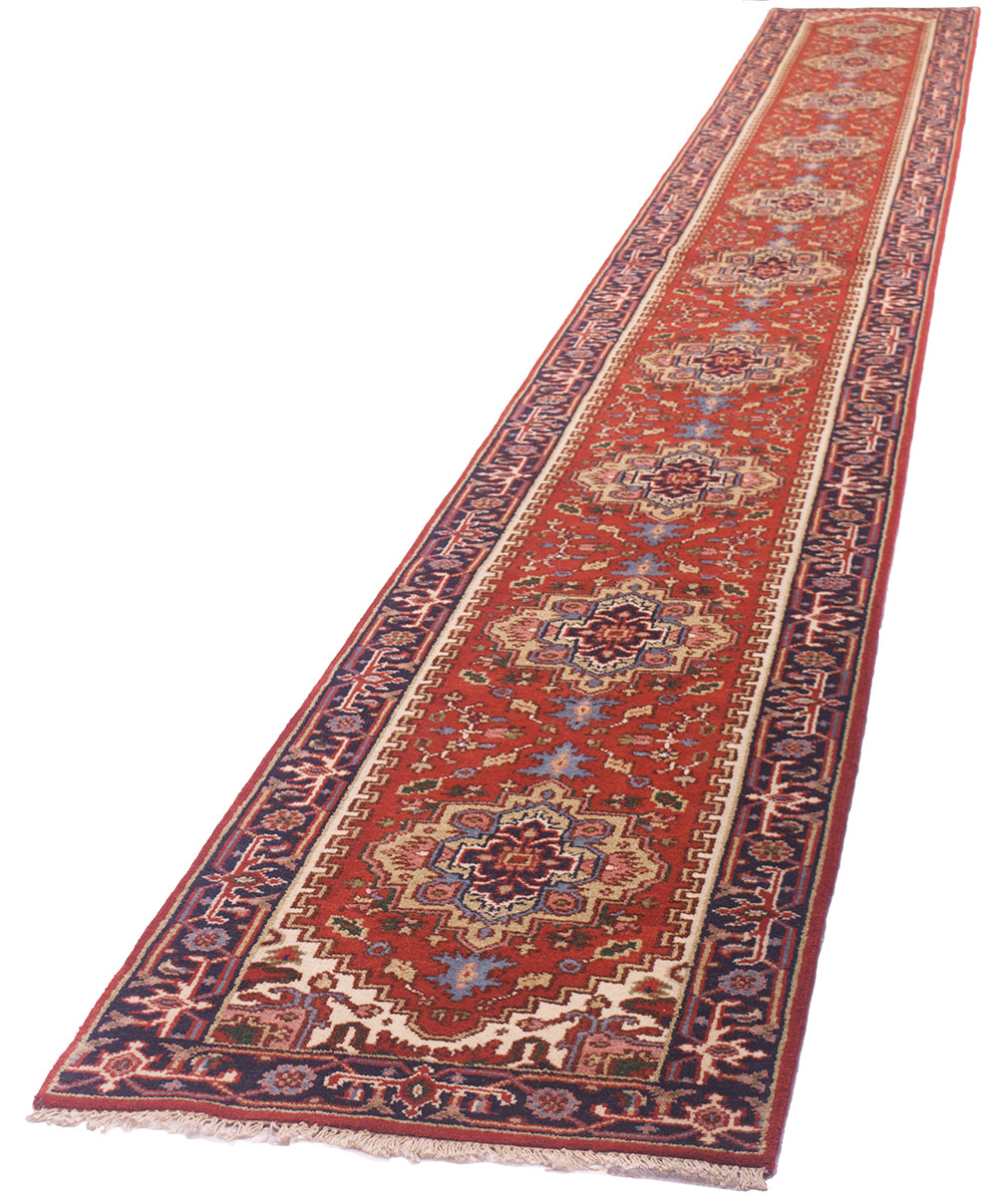 A 24 foot long Indo-Heriz runner shown diagonally on a plain white background. The rug has a warm red-orange field with repeating central medallions and ornate borders in cream, navy, burgundy, and small blue accents.