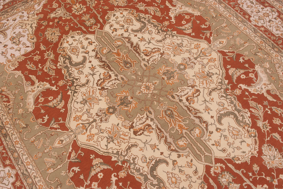 A close-up of an ornate traditional patterned rug with a large central medallion. The design features a warm rust red background with beige, gray, and orange floral and scroll motifs.