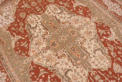 A close-up of an ornate traditional patterned rug with a large central medallion. The design features a warm rust red background with beige, gray, and orange floral and scroll motifs.