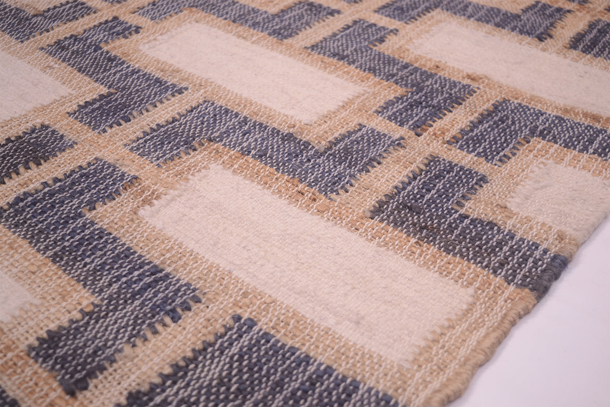 A close-up of a woven rug or textile with a geometric block pattern in beige, cream, and muted blue-gray, showing the texture and corner detail.