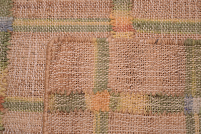 A close-up of a rustic woven textile or rug with a patchwork design, featuring natural tan burlap-like sections and stitched blocks in muted green, yellow, and orange.