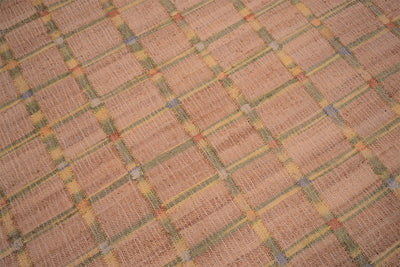 A close-up of a woven textile or rug with a tan and beige base, overlaid with a repeating plaid pattern in muted yellow, olive green, and small blue accents.