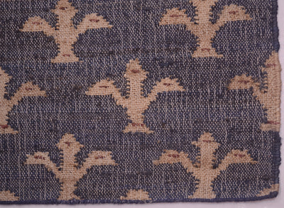 A close-up of a woven textile showing a repeating stylized fleur-de-lys pattern in tan/beige on a dark blue background.