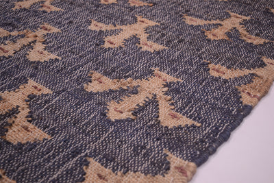 A close-up of a woven textile with a repeating fluer-de-lyspattern in beige and dark charcoal gray, showing the texture and edge of the rug