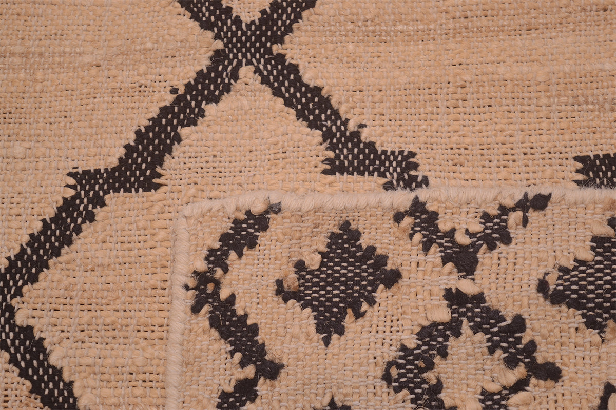 Close-up of a woven rug or textile with a beige natural-fiber background and a black geometric pattern, including a visible corner with a white binding edge.