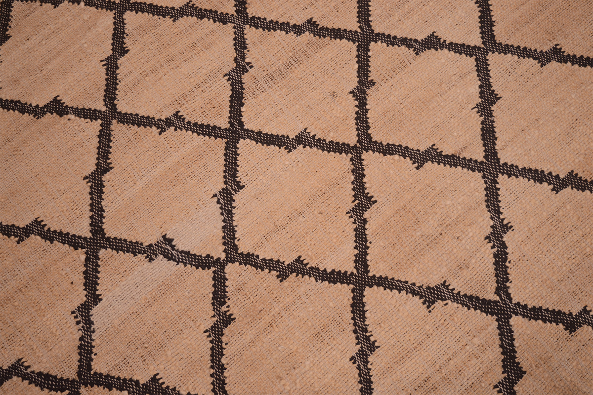 Close-up of a woven rug or textile with a tan-beige background and a dark charcoal-brown geometric diamond grid pattern.