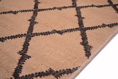 Close-up of a woven rug or textile in warm beige with a dark charcoal geometric diamond pattern, shown against a white background.