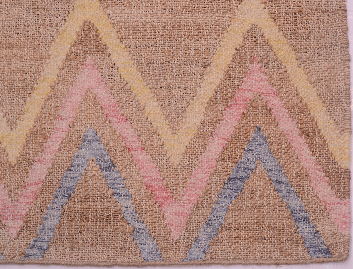 A close-up of a woven jute kilim with a zigzag chevron pattern in muted pink, dusty blue, pale yellow, and beige tone.
