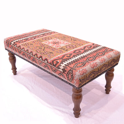 Bench Stool upholstered with old Turkish Kilim with centralised square medallion with muted colours of ligth olive green, rose pink/terracotta dark soft brown, cream. Mid brown wooden legs. Shown on a white background.