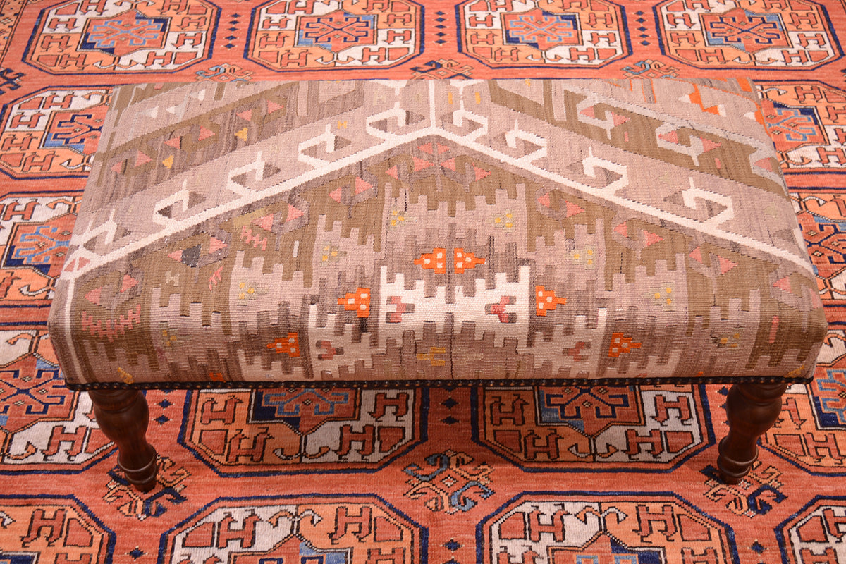Kilim bench footstool upholstered wtih softened colours of vintage turkish kilim. Wooden legs. Vintage kilim textile containing colours of cream, orange, coral pink, beige and light olive. Hints of golden ochre visible. Placed on a terracotta carpet with Afghhan Ersari Fil-pai design. 