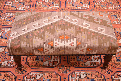 Kilim bench footstool upholstered wtih softened colours of vintage turkish kilim. Wooden legs. Vintage kilim textile containing colours of cream, orange, coral pink, beige and light olive. Hints of golden ochre visible. Placed on a terracotta carpet with Afghhan Ersari Fil-pai design. 