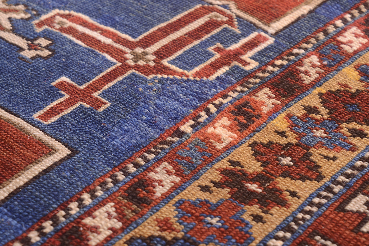 Close-up of a handwoven antique  Caucasian Karagashli rug showing a blue field with a red and cream geometric motif and a patterned border in red, beige, and blue.