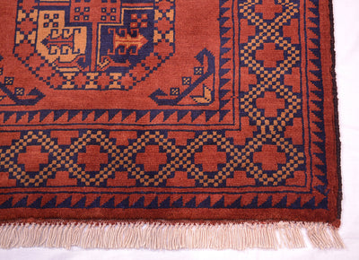 A close-up of a patterned handmade Afghan rug corner showing a rust red field, navy blue geometric border, and cream fringe.