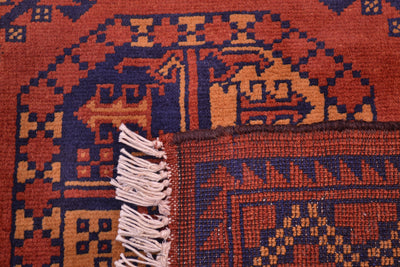 Close-up of the back corner underside to reveal the hand-knotted back of a patterned Afghan Kargahi rug showing a rust red and navy blue geometric design with cream fringe on the edge.