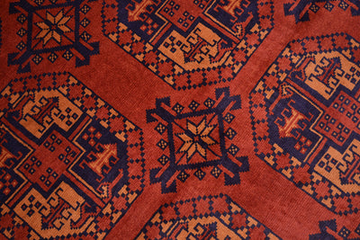 A close-up of a patterned Afghan Kargahi rug with a rust red base, dark navy geometric motifs, and soft orange accents.