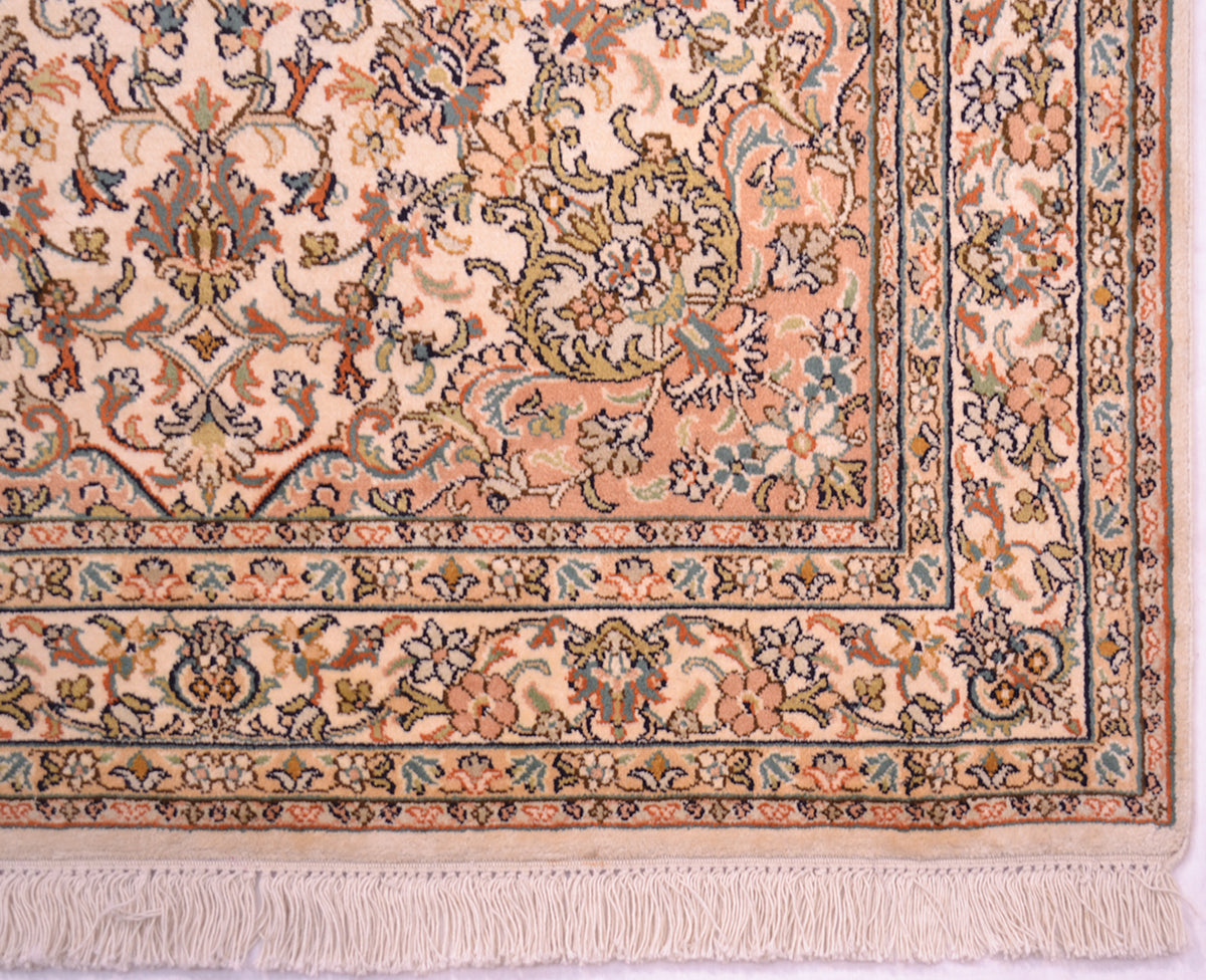 A close-up of a traditional patterned rug corner showing a cream and peach field with floral motifs and a decorative border in beige, blue, green, and rust tones, finished with a cotton fringe.