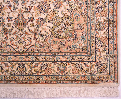 A close-up of a traditional patterned rug corner showing a cream and peach field with floral motifs and a decorative border in beige, blue, green, and rust tones, finished with a cotton fringe.