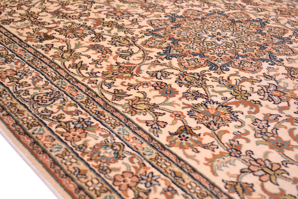 A close-up of an ornate silk area rug with a cream background and intricate floral motifs in navy blue, rust orange, and olive green, showing the detailed woven texture and corner border.