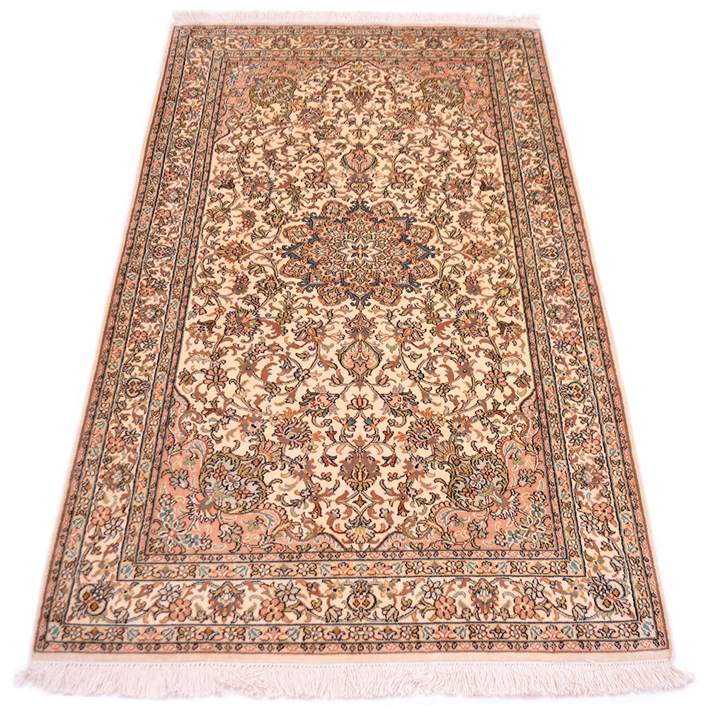 A rectangular silk area rug with a traditional floral and medallion pattern, featuring a beige and peach base with brown, blue, and rust accents, finished with white fringe on the ends.