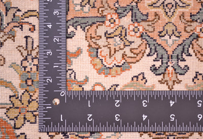 Close-up of a silk rug with a dark ruler placed vertically and horizontally to show scale, featuring a beige, orange, gray, and navy ornamental floral design.