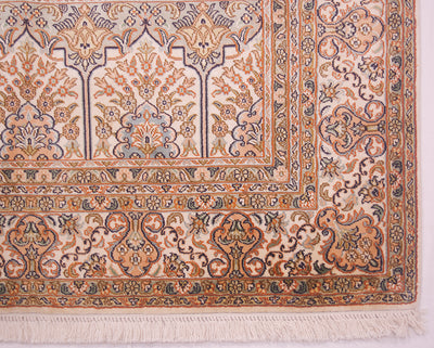 A close-up of an ornate Kashmir silk area rug corner, showing intricate floral patterns in beige, rust orange, and navy blue with white fringe.