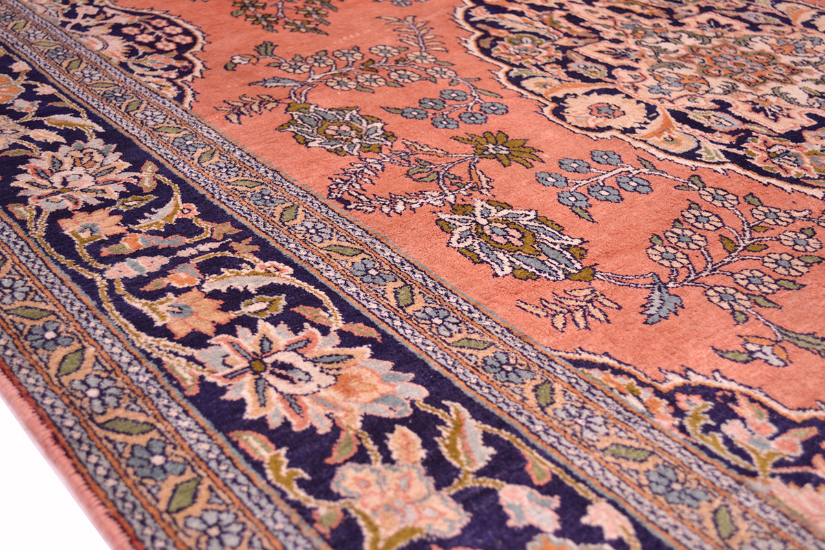 A close-up of an ornate pattern silk Kashmir rug with a pink field, dark navy borders, and floral motifs in beige, green, and blue.