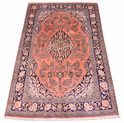 A silk Kashmir rug with a coral pink field, navy blue borders, and intricate floral motifs in beige, green, and blue, shown flat against a white background.