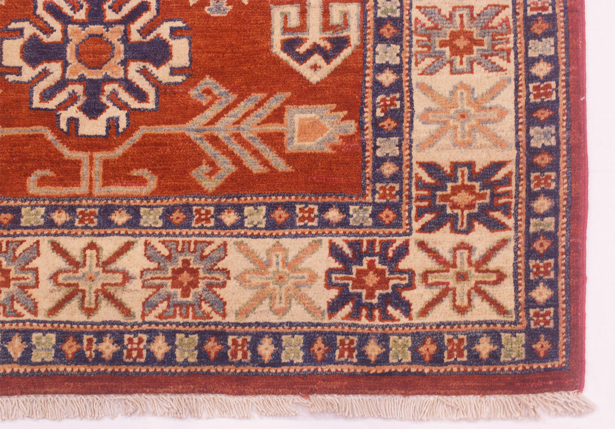 Kazak rug corner close-up with cream-and-blue border, rust-red motifs, short fringe