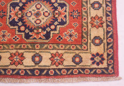A close-up of a traditional small size handmade Kazak Mat corner showing a red field, beige border, navy details, and fringed edge.