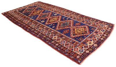Angled view of Antique kazak  long rug with deep blue field. Handknotted and handmade with white highlights, black, reds, blues and yellow golds with multiple borders and geometric devices. Shown on a white background. Woven on wool warps  with wool pile.