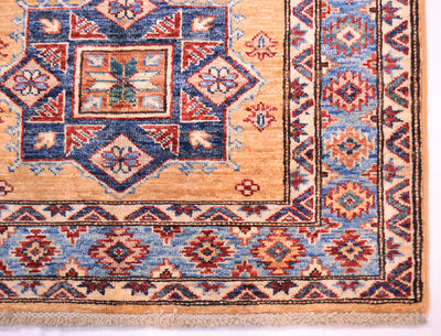 Close-up of a patterned Afghan Kazak rug corner showing a yellow-amber field, blue and red geometric medallion motifs, and a decorative border with fringe along the bottom edge.