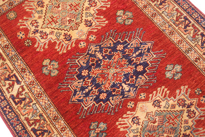A close-up of a traditional patterned super Kazak rug with a dominant red field, a large navy blue central medallion, and beige geometric borders.