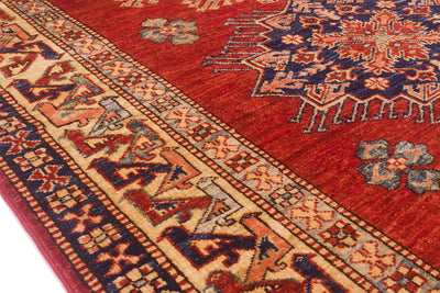 A close-up of a traditional Afghan Kazak patterned rug with a deep rust-red field, beige and navy borders, and blue floral motifs.