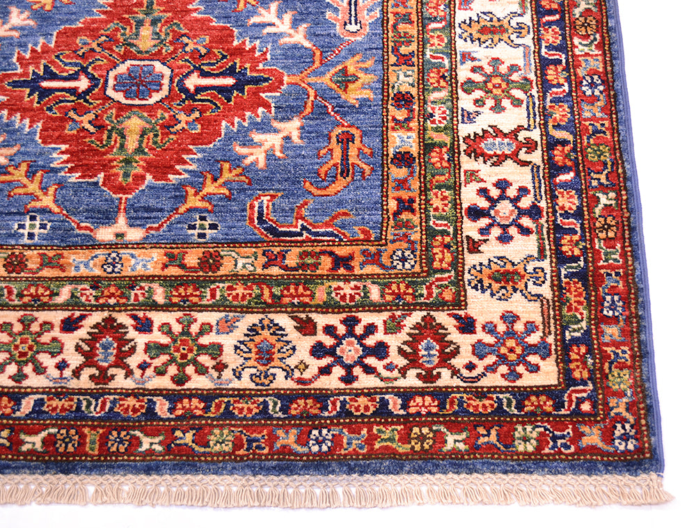 A close-up of an ornate patterned fine Kazak Afghan rug, corner showing a blue field, cream border, and red, green, and orange floral motifs with fringe along one edge.