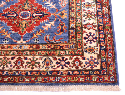 A close-up of an ornate patterned fine Kazak Afghan rug, corner showing a blue field, cream border, and red, green, and orange floral motifs with fringe along one edge.