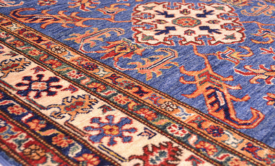 A close-up of a traditional Afghan Kazak patterned rug with a medium blue field, cream borders, and orange, red, and navy floral motifs.
