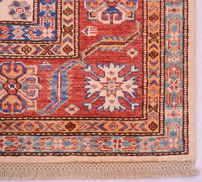 A close-up of a traditional handwoven fine Afghan Kazak carpet corner showing a red main border with cream, blue, and brown geometric and floral motifs, finished with a cream fringe.