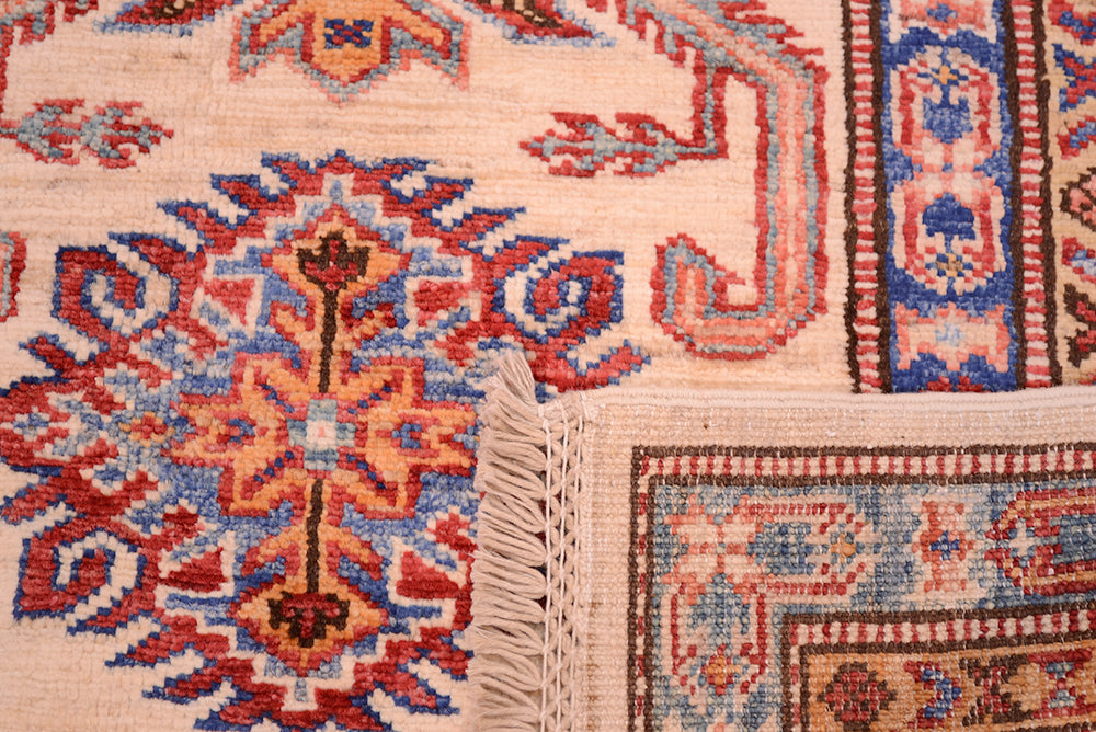 Close-up of the corner turned up of hand-knotted fine Afghan Kazak patterned rug corner showing cream, red, blue, and orange woven textile with fringe and detailed geometric motifs.
