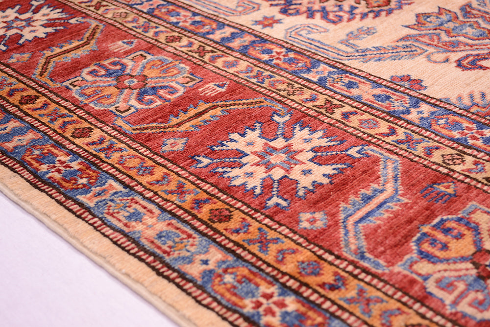 A close-up of a traditional patterned fine Afghan Kazak hand-knotted carpet with a red border, cream field, and blue and orange motifs, shown on a white surface.