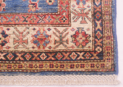 A close-up of a traditional patterned Kazak rug corner showing cream, blue, red, and dark navy woven motifs with fringe along the bottom edge.