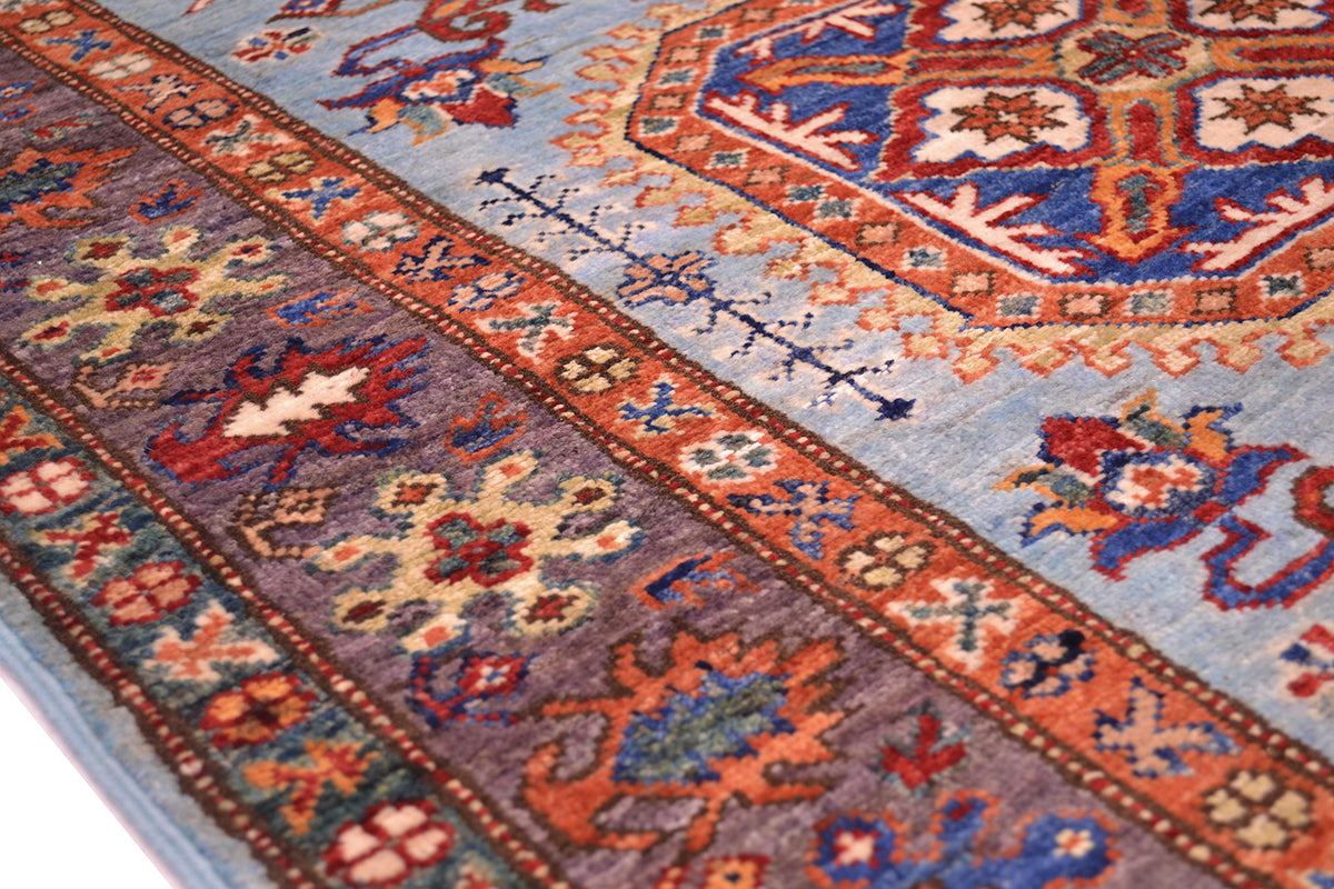A close-up of a traditional patterned Afghan Kazak rug with a light blue field, ornate medallion motifs, and a aubergine border featuring floral and geometric designs in rust, blue, cream, and red.