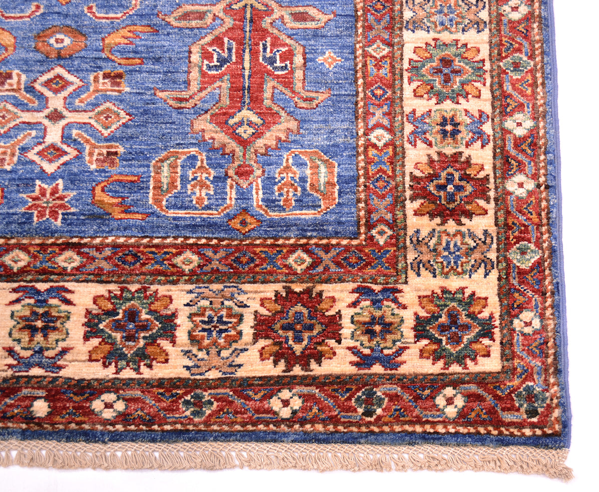 A close-up of a traditional patterned rug corner showing a blue field with ornate red, beige, and navy motifs, a decorative border, and a short fringe.