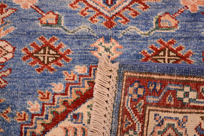 Close-up of the hand-knotted back with corner turned up of an Afghan Fine Kazak small rug showing a blue field with red, beige, and cream geometric motifs and a cream fringe.
