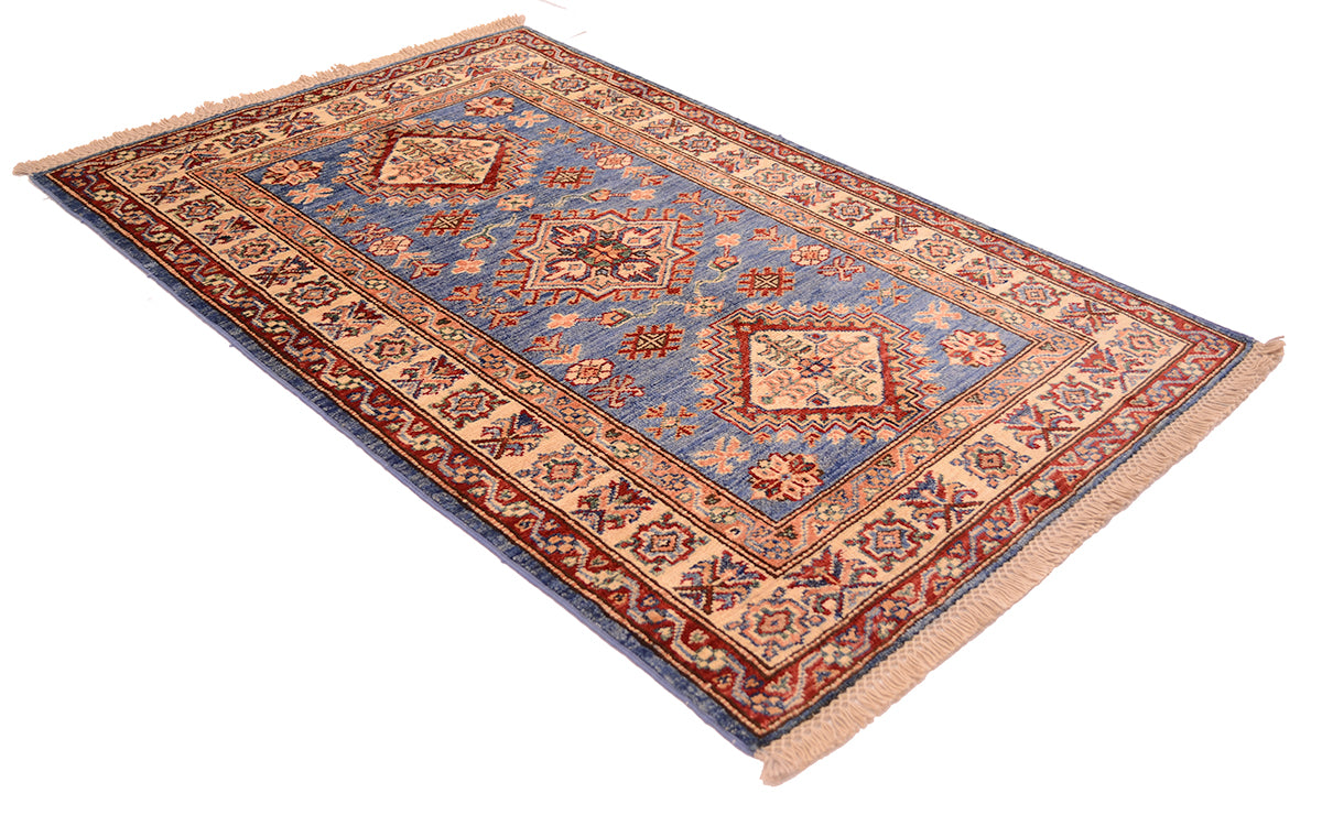 A rectangular traditional small Afghan Kazak rug with a blue field, red and beige borders, and fringed edges, shown at an angle on a white background.