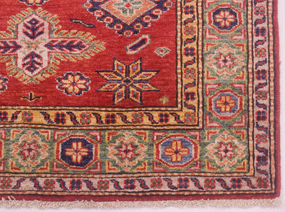 A close-up of a traditional patterned Afghan Kazak rug corner showing a red field with geometric medallions and a multicolored border in blue, green, beige, and orange.