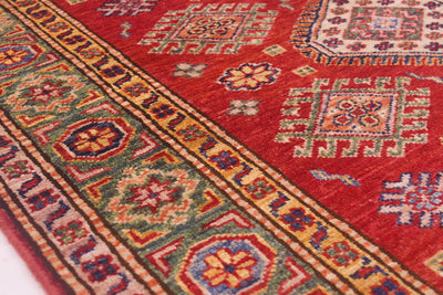 A close-up of a traditional patterned fine Kazak handmade rug with a dominant red field, featuring geometric medallions and a detailed border in beige, green, blue, and orange.